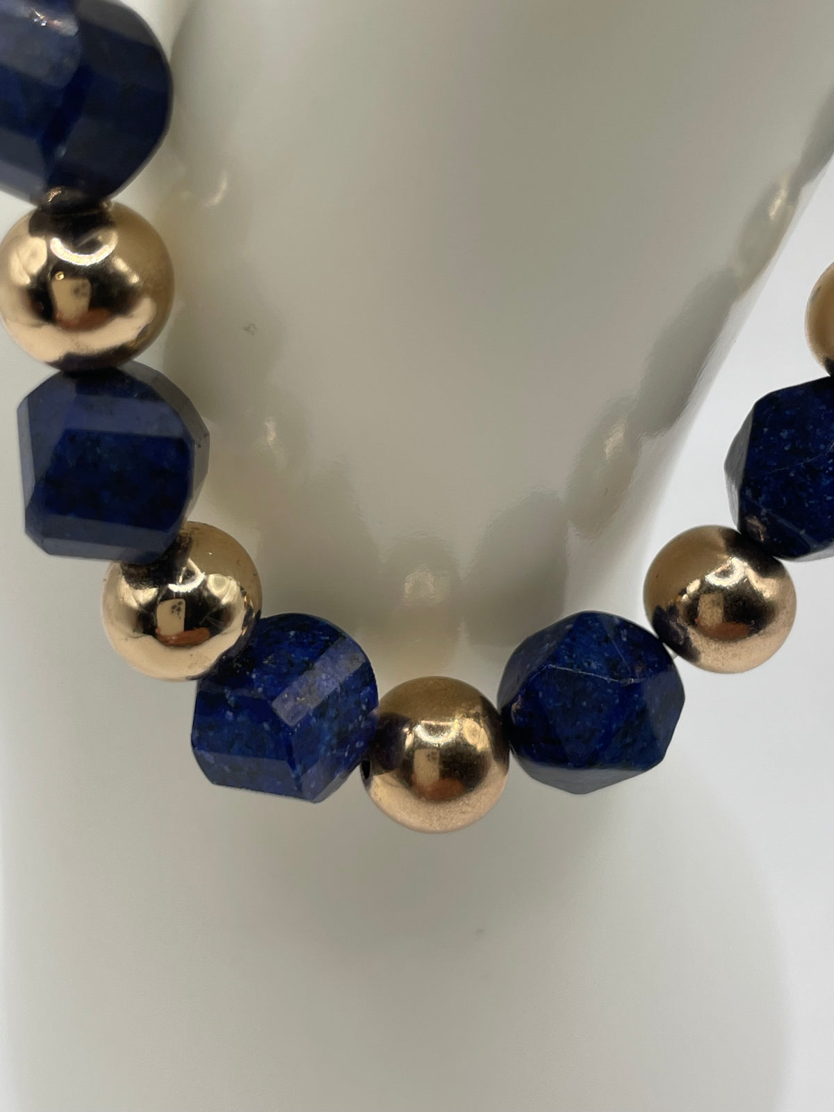 A beaded bracelet featuring faceted dark blue lapis lazuli stones and polished metallic gold round beads, displayed on a white cylindrical stand.