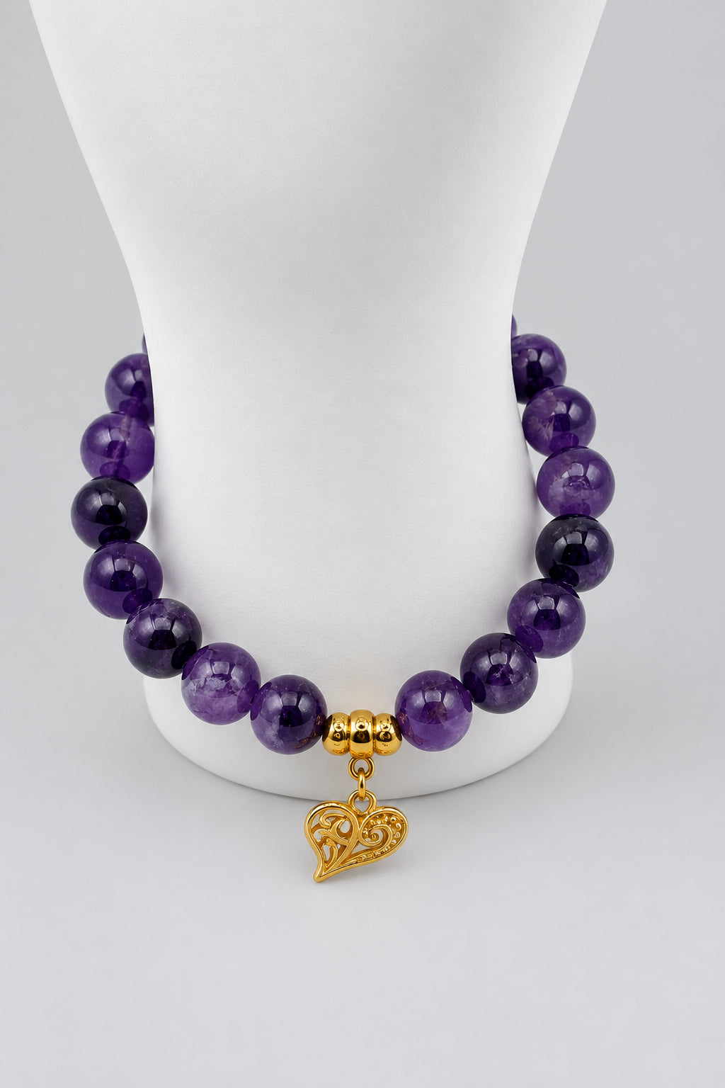 A beaded bracelet made of polished purple gemstone beads with a small gold heart-shaped charm, displayed on a white mannequin stand against a white background.