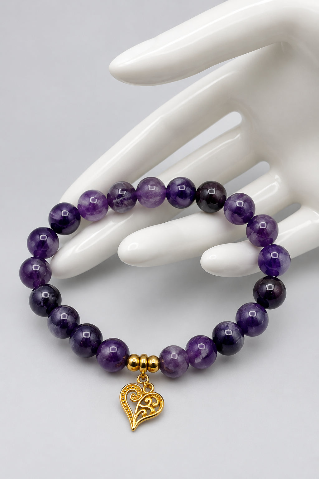 A beaded bracelet made of round purple gemstone beads with a small gold heart charm, displayed on a white ceramic mannequin hand against a light gray background.