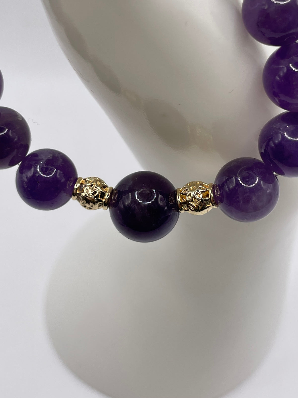 A close-up of a beaded bracelet with large polished purple beads and two ornate gold spacer beads, displayed on a white mannequin stand.