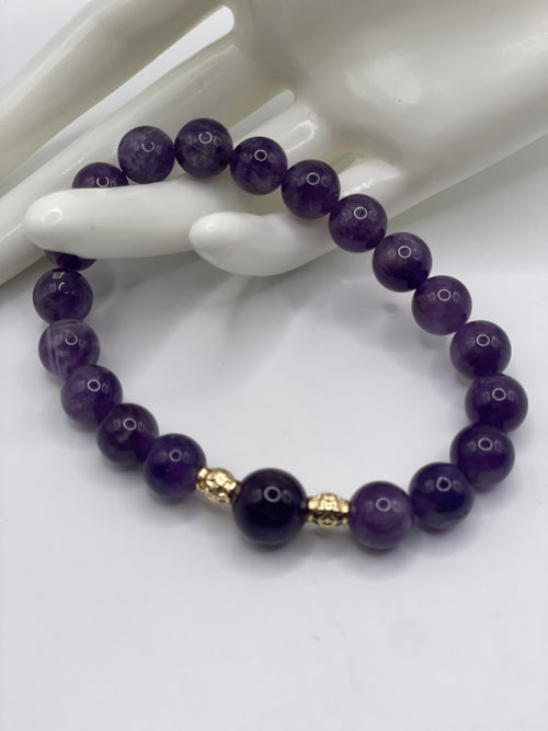 A beaded bracelet made of round purple gemstone beads with two small gold-toned spacer beads, displayed on a white mannequin hand against a white background.