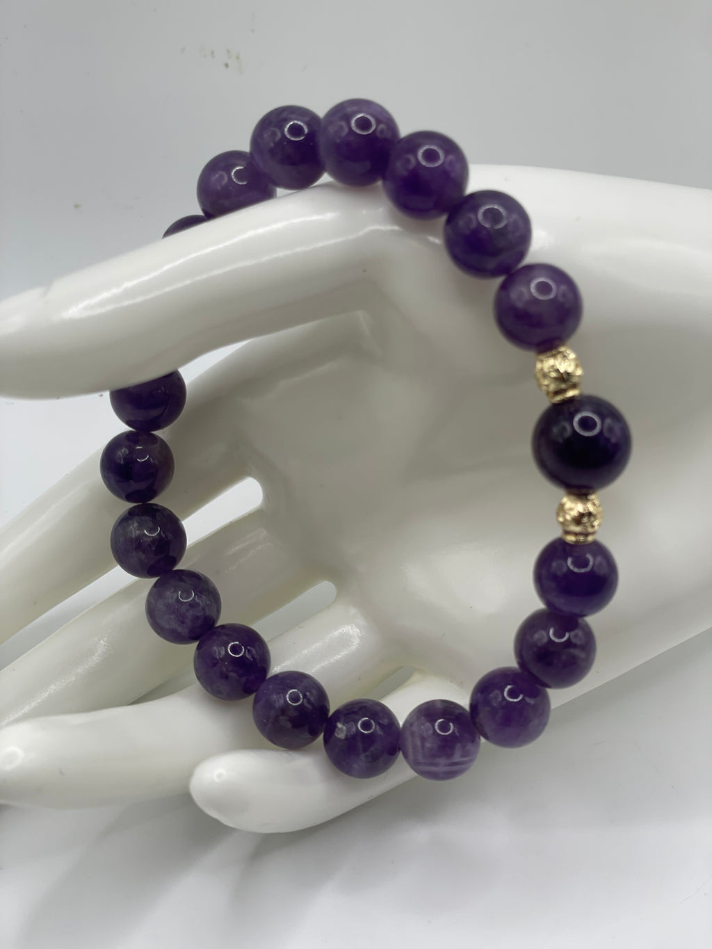 A beaded bracelet made of polished purple gemstone beads with two small gold-toned spacer beads, displayed on a white ceramic hand-shaped stand against a light gray background.
