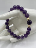 A beaded bracelet made of polished purple gemstone beads with two small gold-toned spacer beads, displayed on a white ceramic hand-shaped stand against a light gray background.
