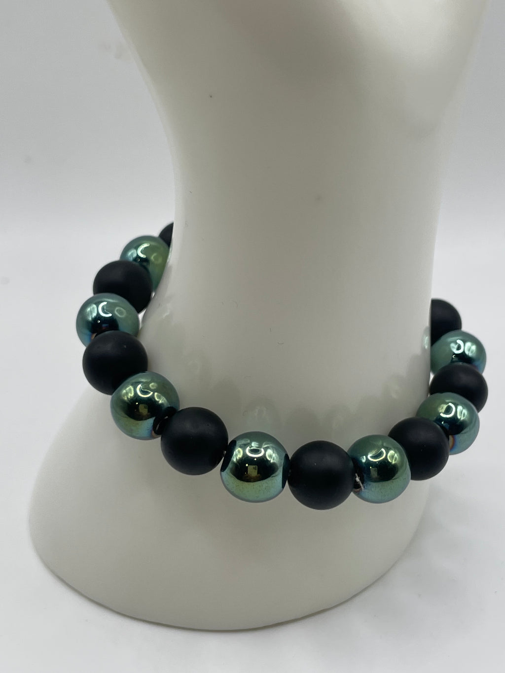 A beaded stretch bracelet with alternating matte black and metallic teal-green beads, displayed on a white mannequin neck.
