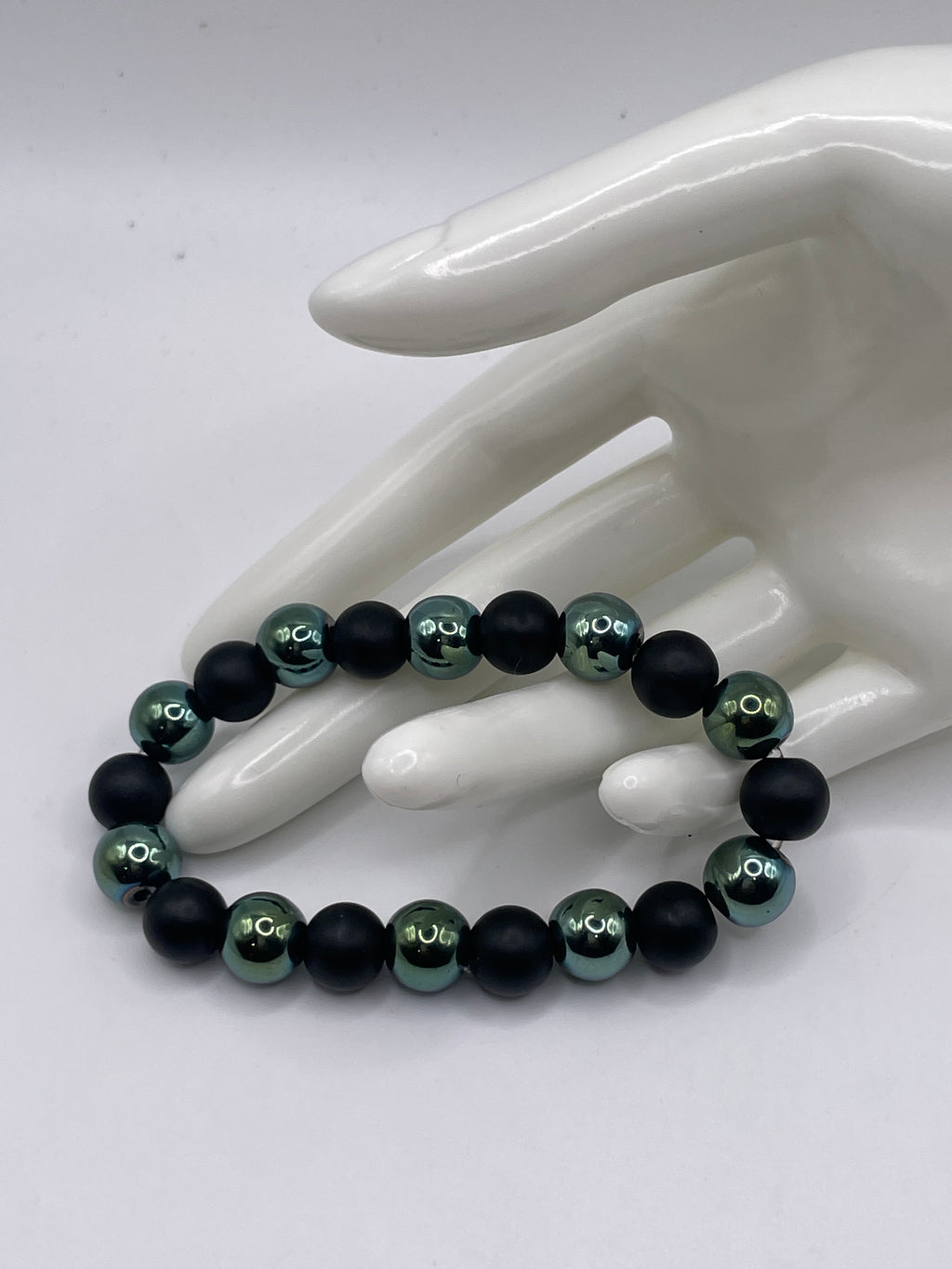 A beaded stretch bracelet with alternating matte black and glossy metallic teal-green beads, displayed on a white ceramic mannequin hand against a light gray background.