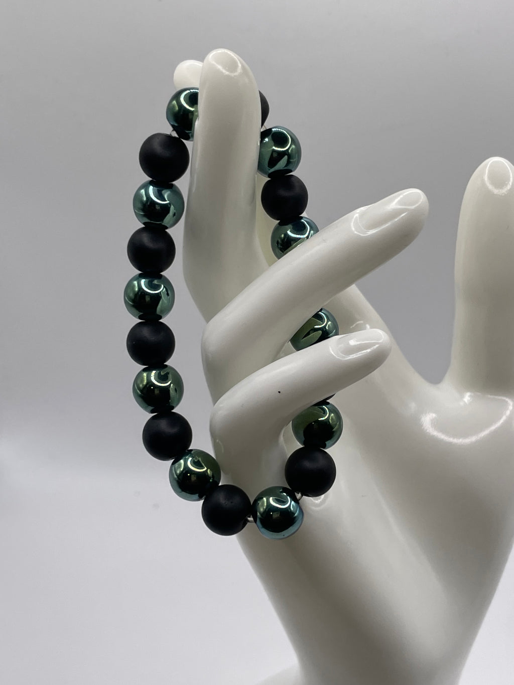 A beaded bracelet displayed on a white ceramic mannequin hand against a light gray studio background. The bracelet features matte black beads alternating with metallic dark green beads that have a reflective finish.