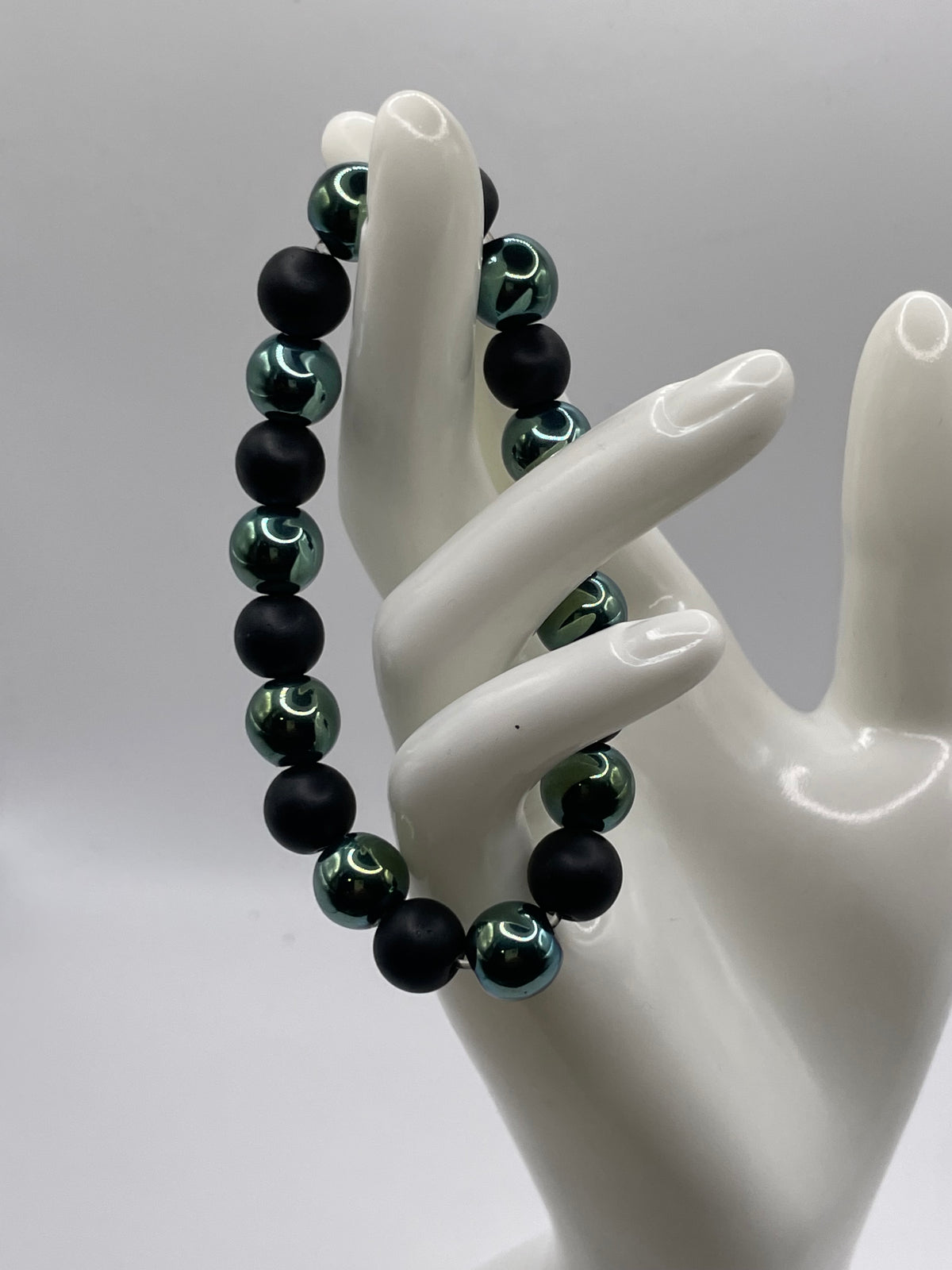 A beaded bracelet displayed on a white ceramic mannequin hand against a light gray studio background. The bracelet features matte black beads alternating with metallic dark green beads that have a reflective finish.