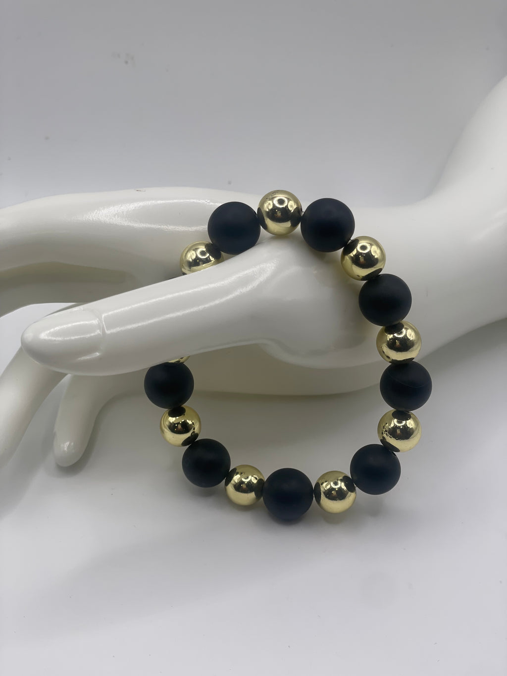 A beaded stretch bracelet with alternating matte black and shiny gold round beads, displayed on a white mannequin hand against a white background.