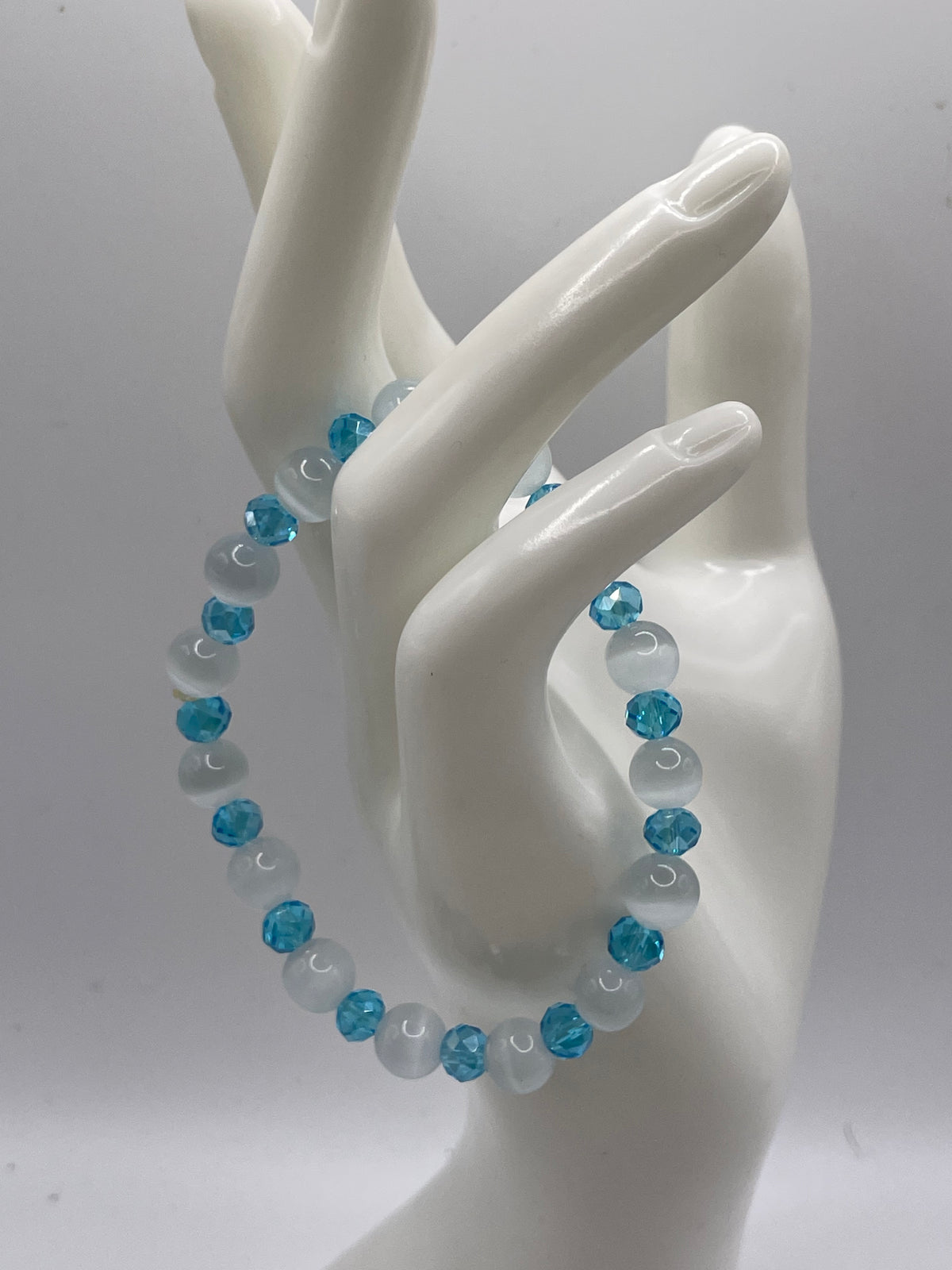 A beaded stretch bracelet with alternating light blue faceted beads and opaque white round beads, displayed on a glossy white ceramic hand mannequin against a light gray background.