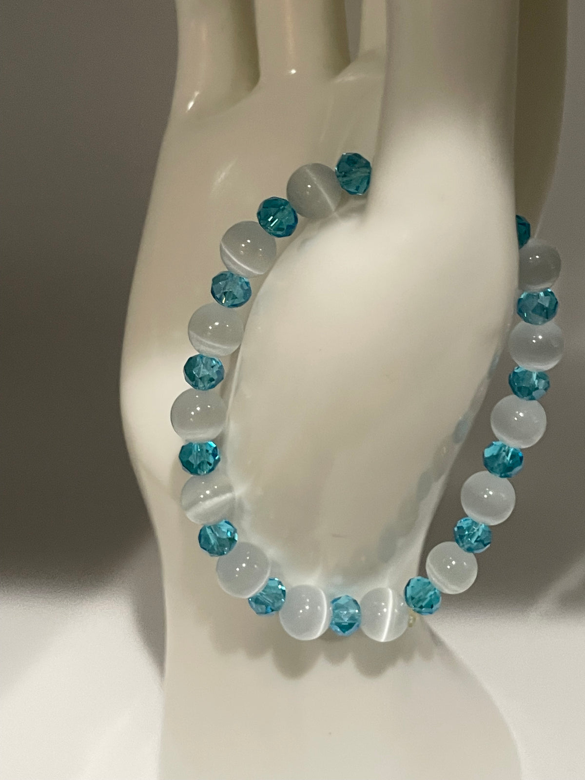 A beaded stretch bracelet with alternating translucent white and faceted light blue beads, displayed on a glossy white ceramic mannequin hand.