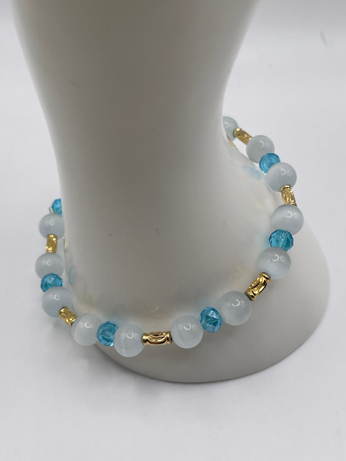 A beaded stretch bracelet displayed on a white mannequin stand. The bracelet features light blue and white beads with small gold spacer accents.