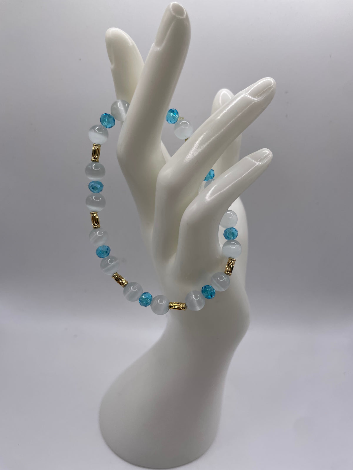 A beaded bracelet with light blue and white beads, accented with small gold spacer beads, displayed on a white ceramic mannequin hand against a light gray background.