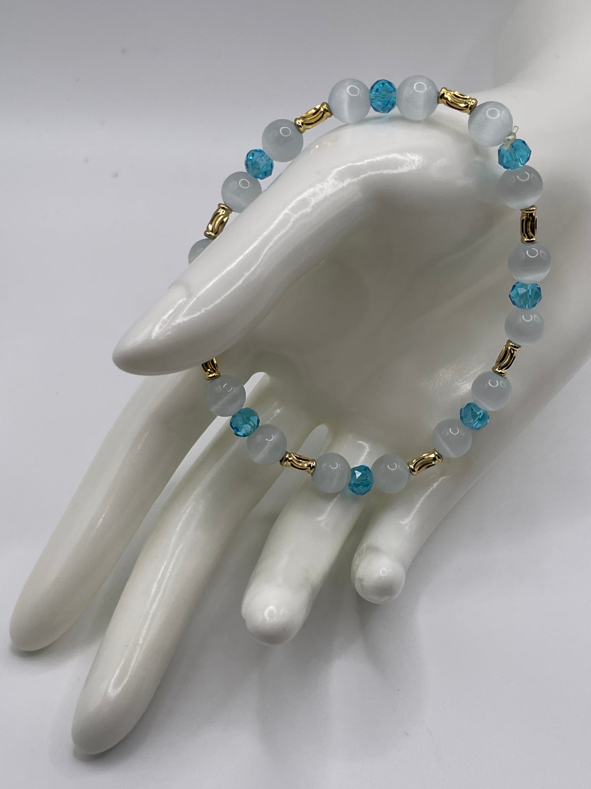 A beaded bracelet displayed on a glossy white ceramic mannequin hand. The bracelet features light blue faceted beads, milky white round beads, and small gold spacer rings.