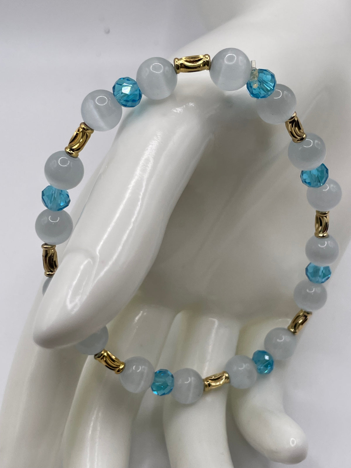A beaded stretch bracelet displayed on a white ceramic mannequin hand. The bracelet features matte white and milky translucent beads, faceted light blue beads, and small gold spacer rings.