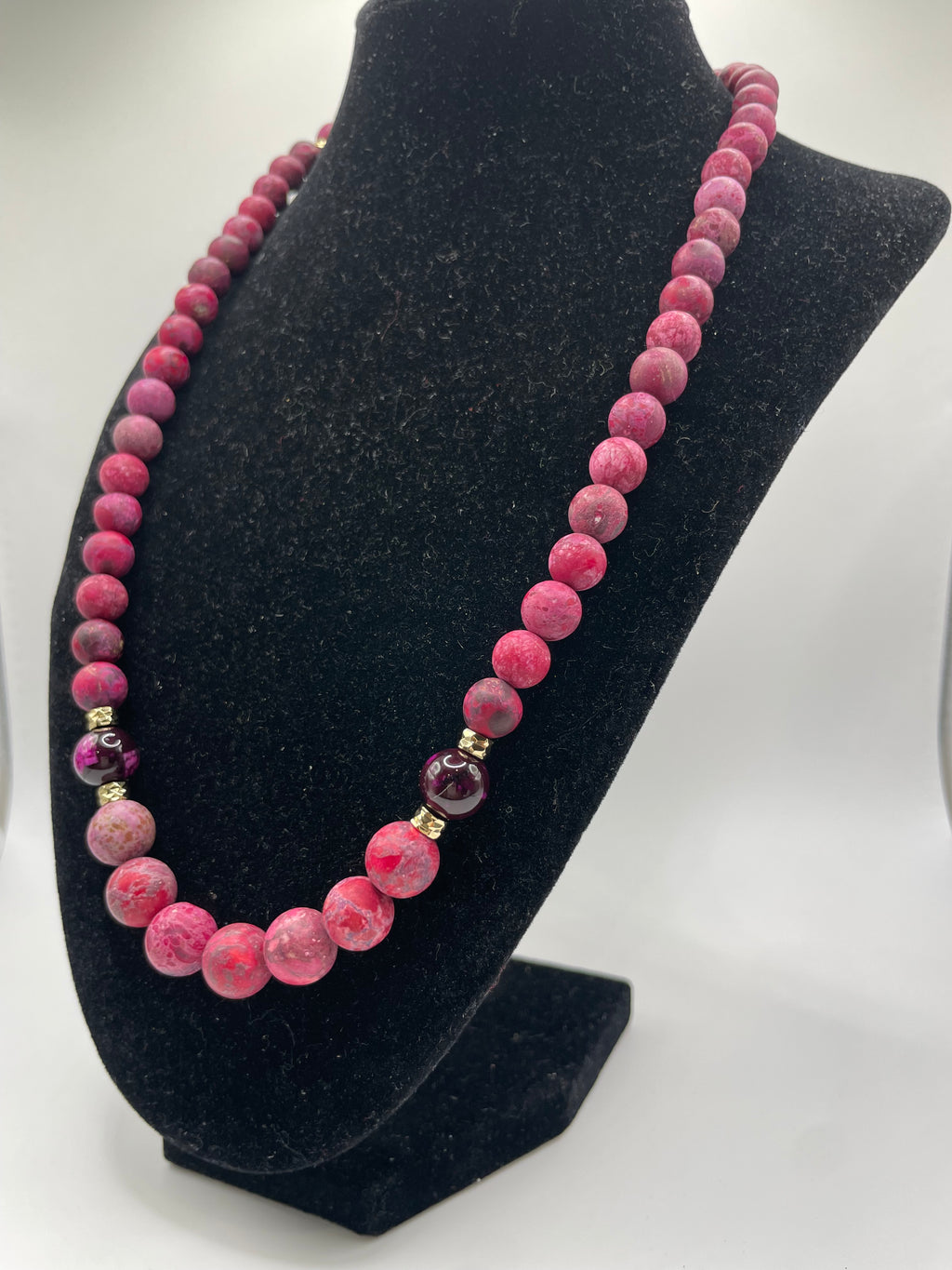 A pink beaded necklace with a gradient of color from light to deep pink, accented by small gold spacer beads and a faceted purple gemstone, displayed on a black velvet jewelry bust.