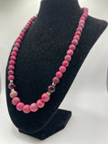 A pink beaded necklace with a gradient of color from light to deep pink, accented by small gold spacer beads and a faceted purple gemstone, displayed on a black velvet jewelry bust.
