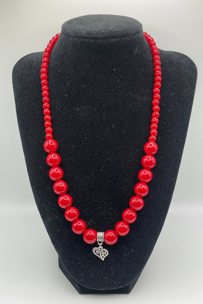 Red beaded necklace with a silver heart charm on a black mannequin bust.