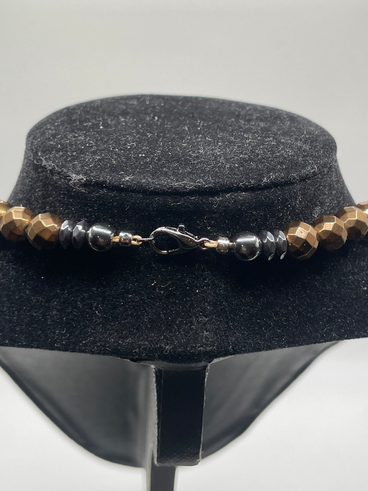 A beaded necklace back view with faceted brown beads, dark metallic spacer beads, and a silver-toned lobster clasp, displayed on a black velvet jewelry bust against a light gray background.
